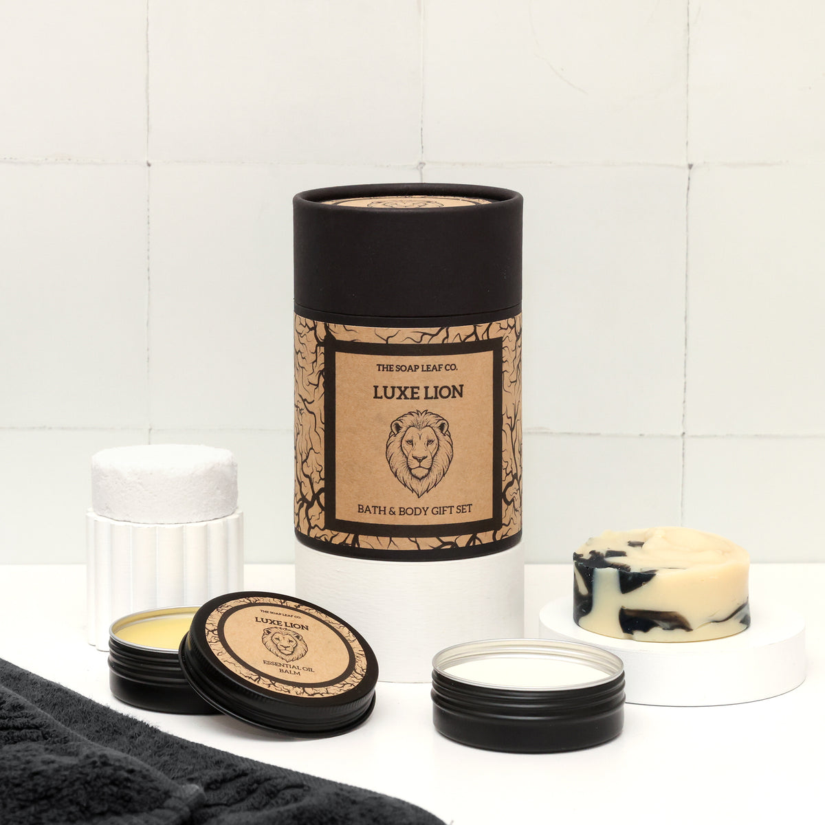 Luxe lion bath and body set on white surface in bathroom with towel. Showing hand soap, shower steamer unwrapped and deodorant and balm in black tins.