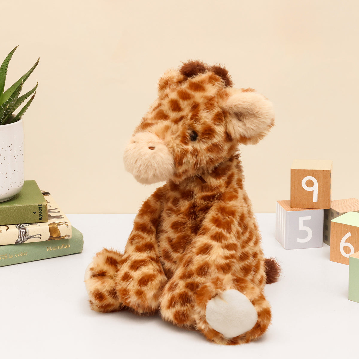 Left side of Giraffe plush made of recycled plastic, sitting on white surface with wooden blocks, books and plant in background