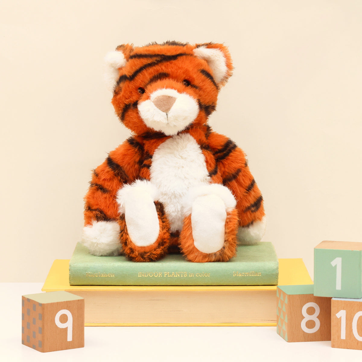 Enchanted world Tiger plush in orange with black stripes and white belly, face and paws. Sitting on books next to wooden blocks on white surface and cream background