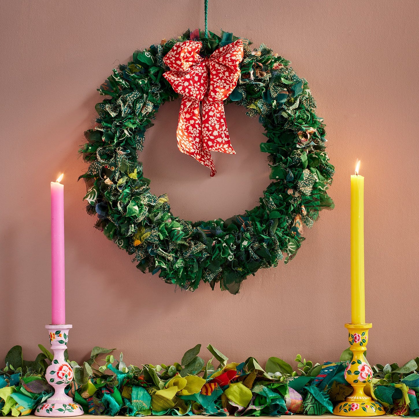 Recycled sari wreath in green with red patterned bow hanging on wall next to 2 lit candle sticks and recycled sari garland in green.