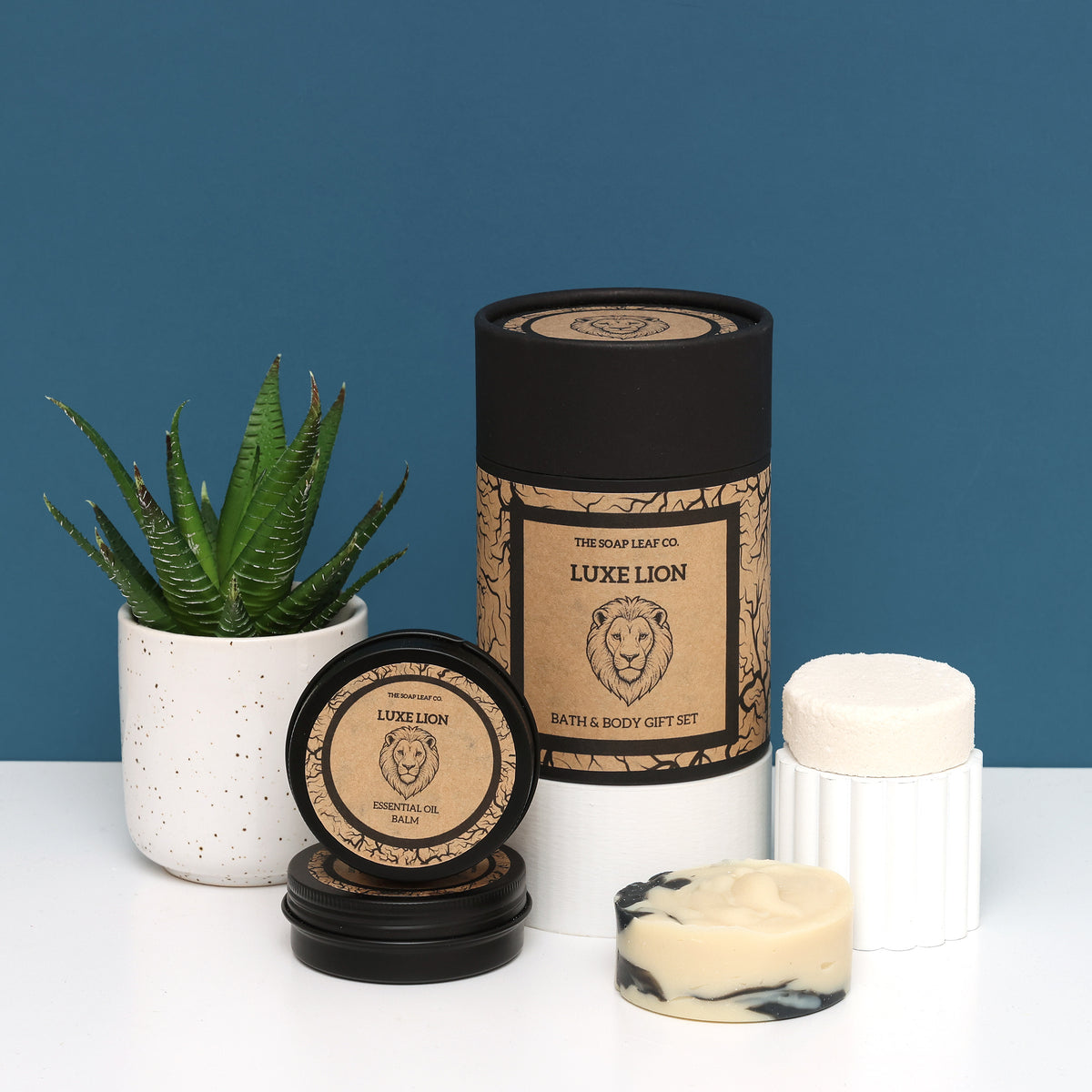 Luxe lion bath and body set on white surface with blue background and plant. Made with essential oils and organic ingredients in a illustrated carboard tube and black tins. 