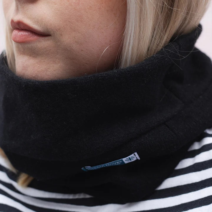 Close-up of a person wearing a black fleece cowl neck warmer over a black and white striped top. A small blue and white brand tag is visible on the neck warmer.