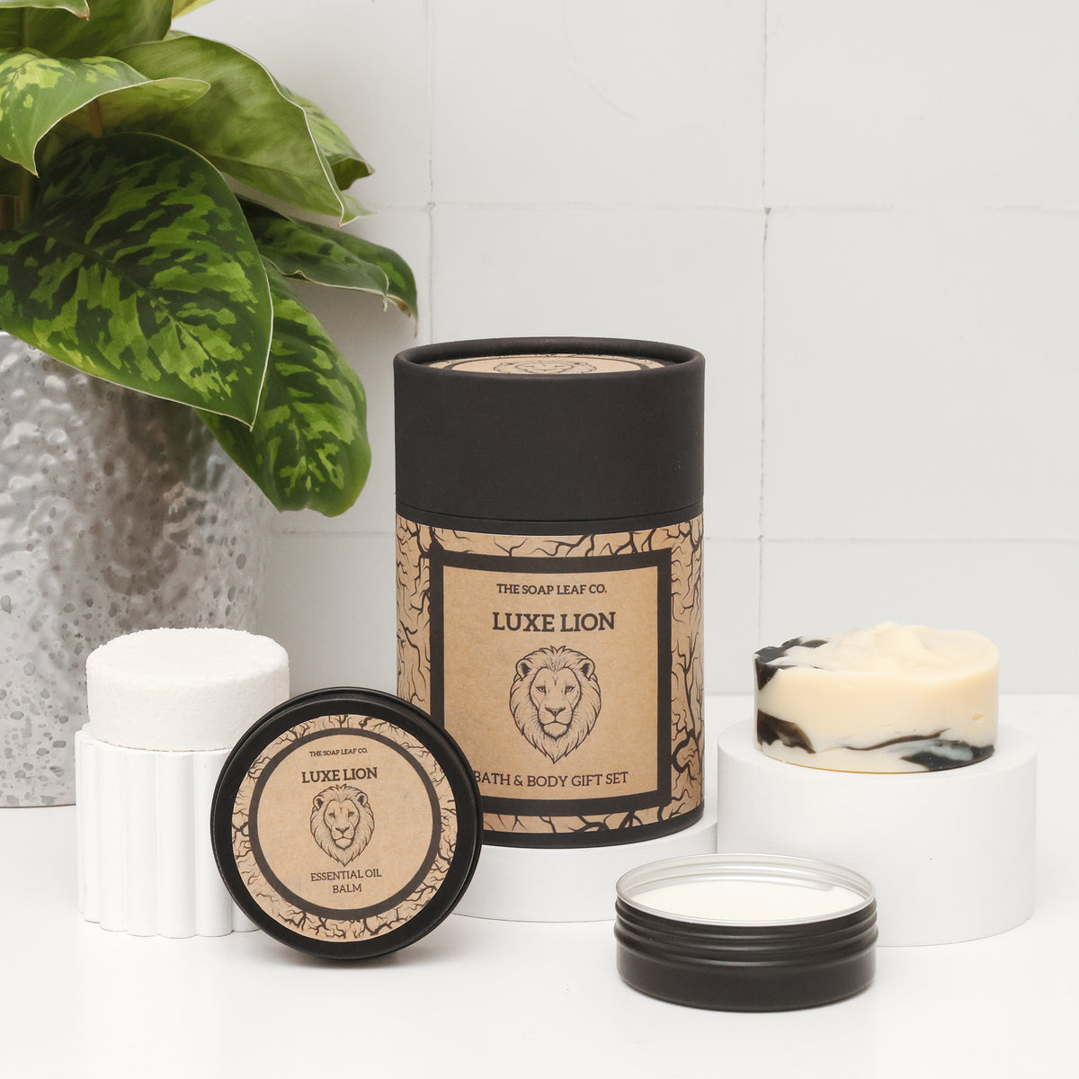 Luxe lion bath and body set on white surface with white tiled background and plant. Showing hand soap, shower steamer, deodorant and face balm