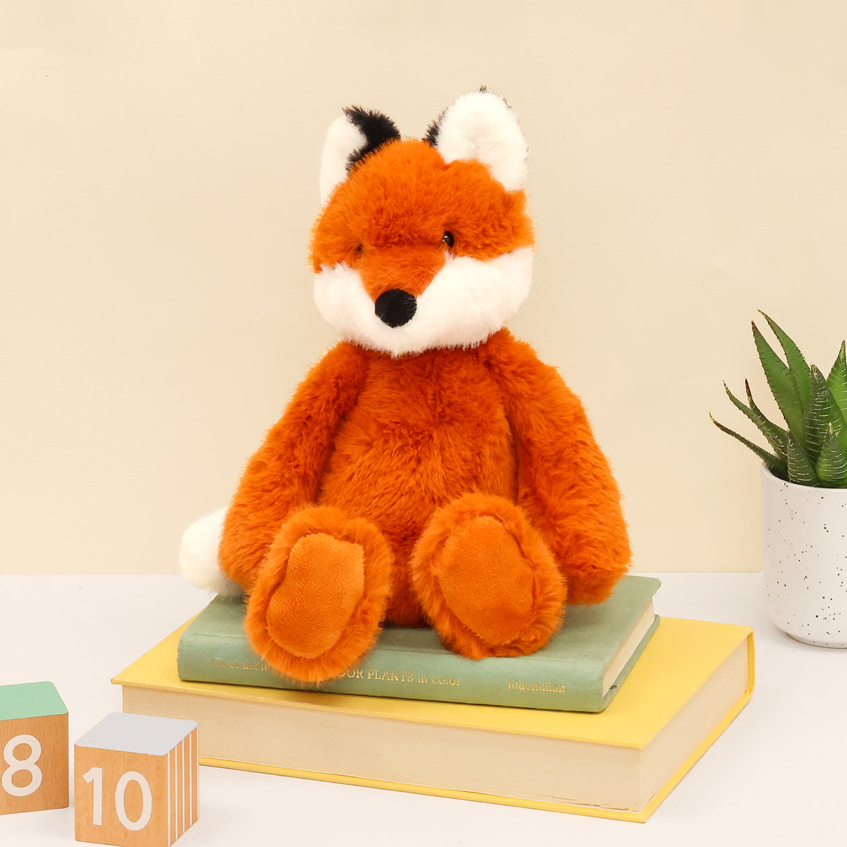 Face on fox plushie made from recycled bottles sitting on books, with wooden blocks and plant in the surface and cream background