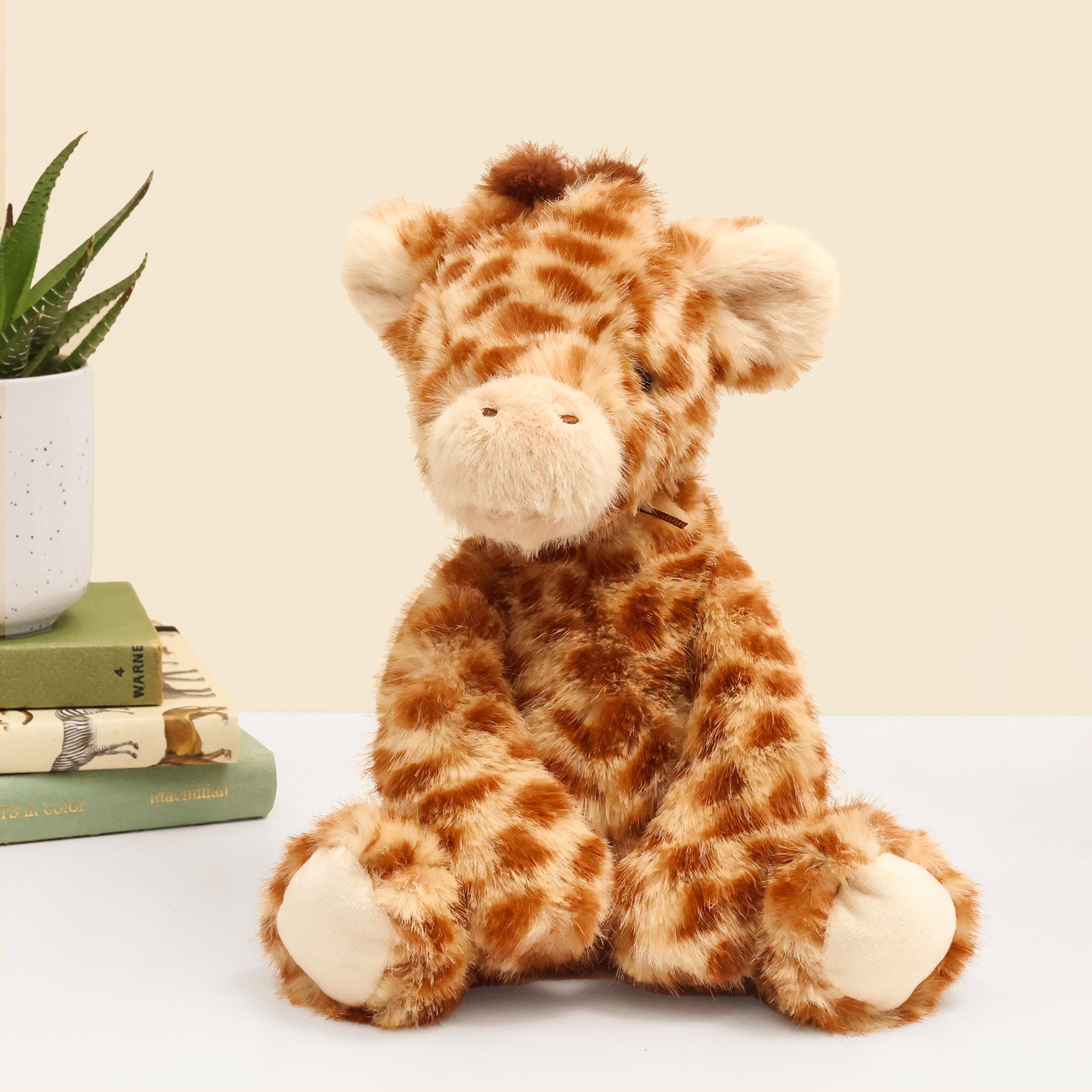 Front facing Super Soft Giraffe plush in beige and brown sitting on white surface with books and plant in background