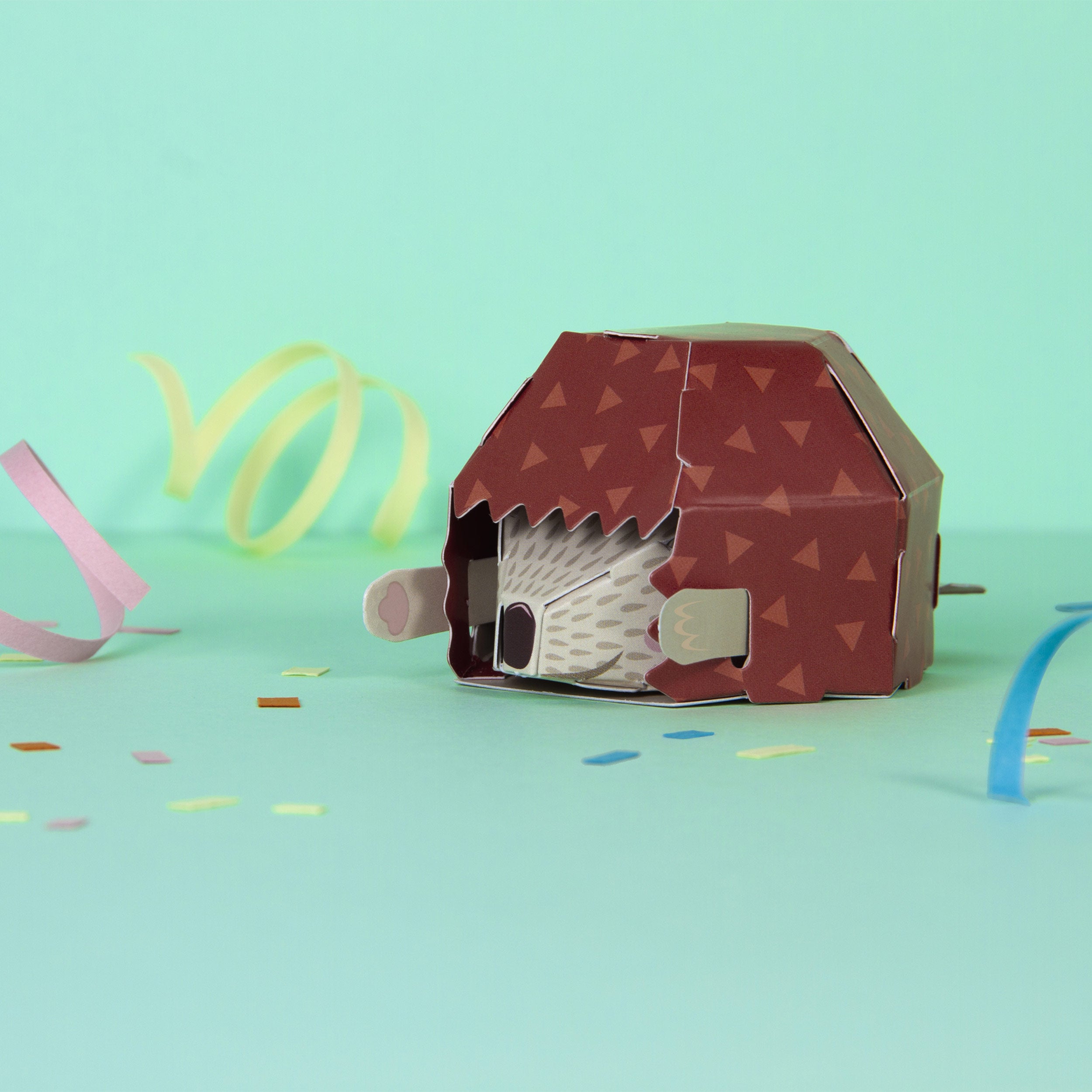 Fully assembled cardboard hedgehog hiding it's head inside it's body on mint green background with party streamers 