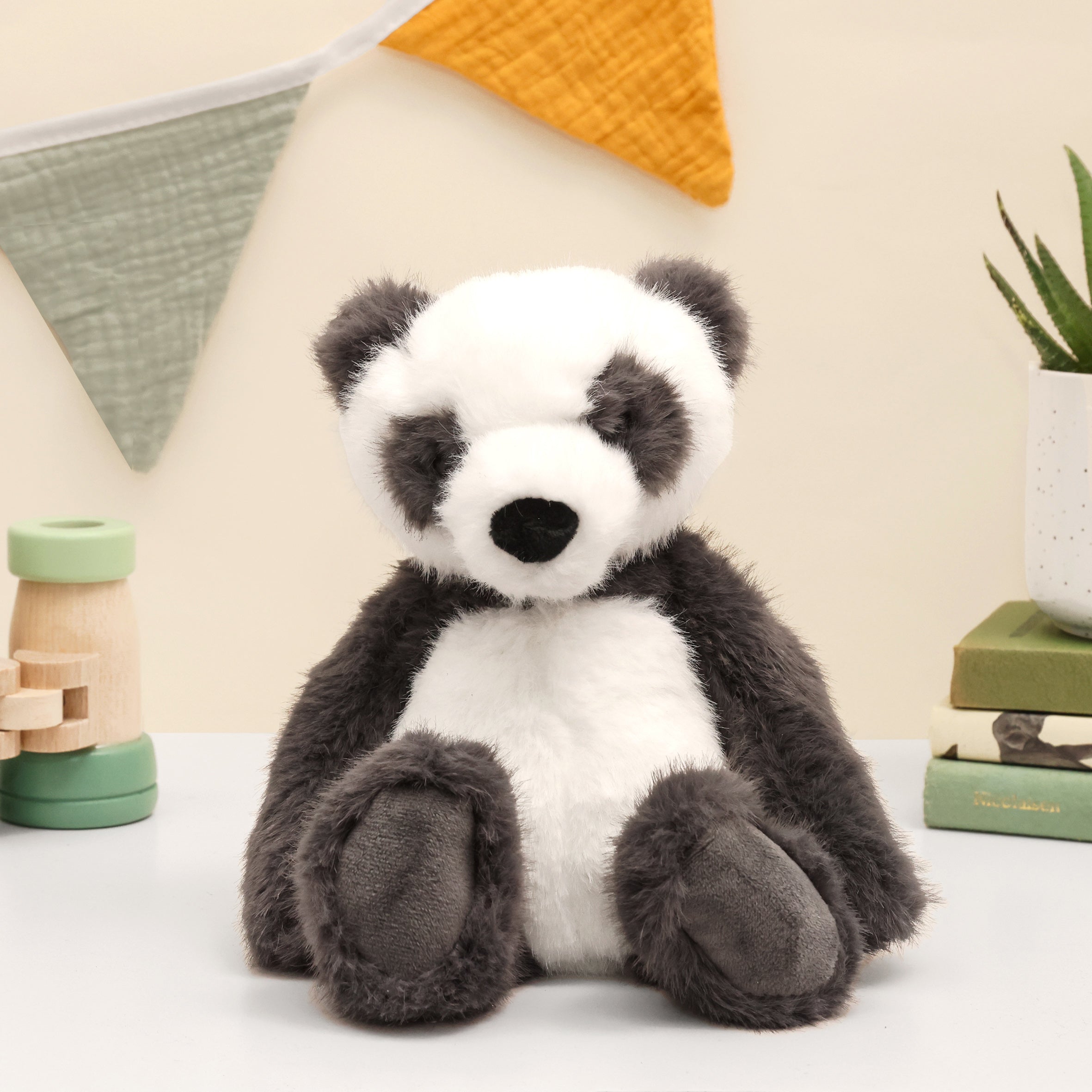 Super fluffy Enchanted world Panda plush in grey and white sitting on white surface with books, plant, wooden binoculars on white surface and bunting on the wall