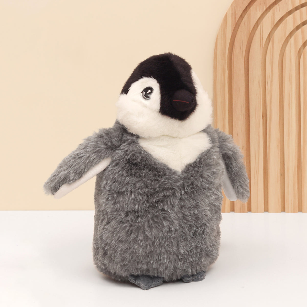 Close up of right side of Baby emperor plush penguin made from recycled bottles in grey standing on white surface with wooden and cream background