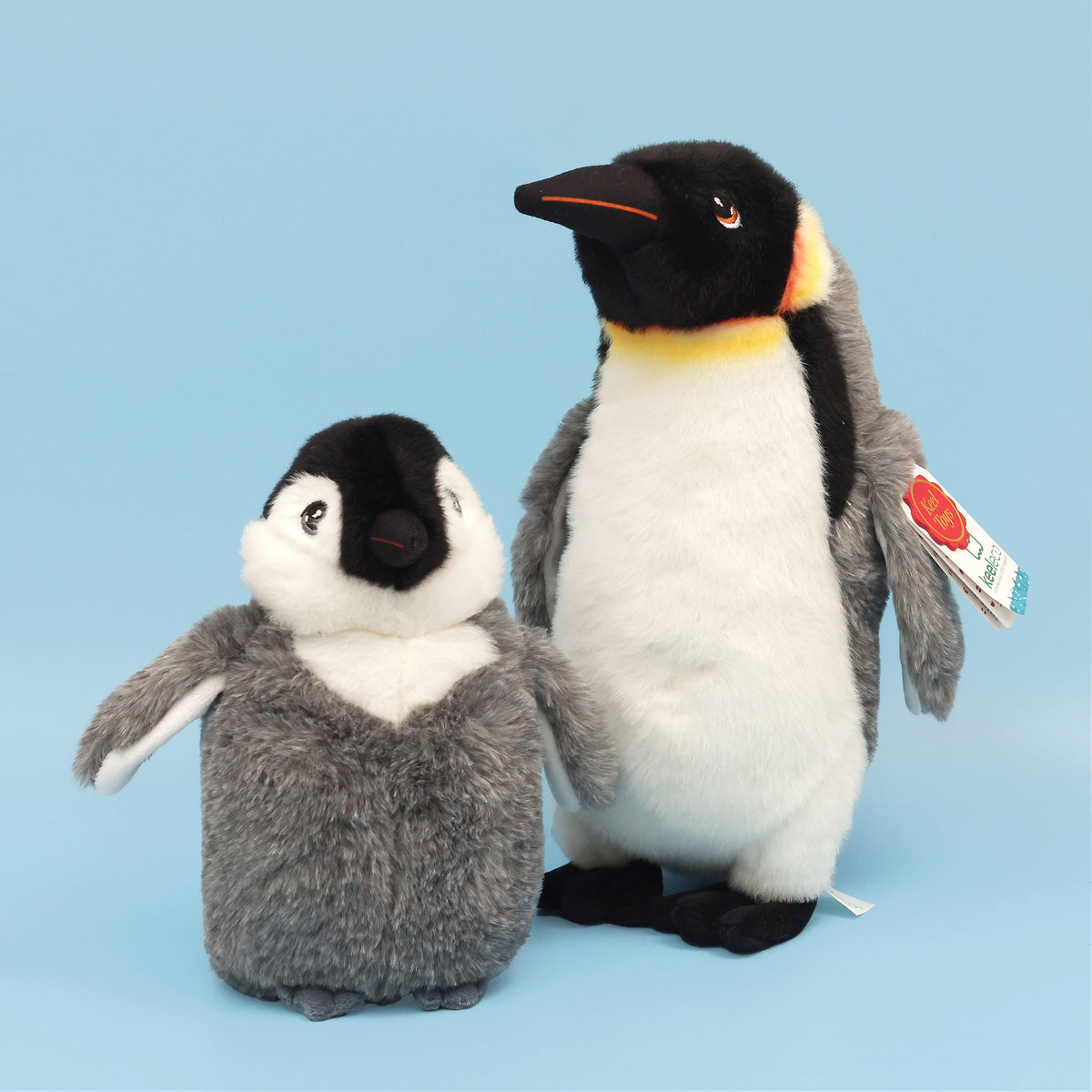 Baby emperor plush penguin with King Emperor penguin plush, standing next to each other on blue background