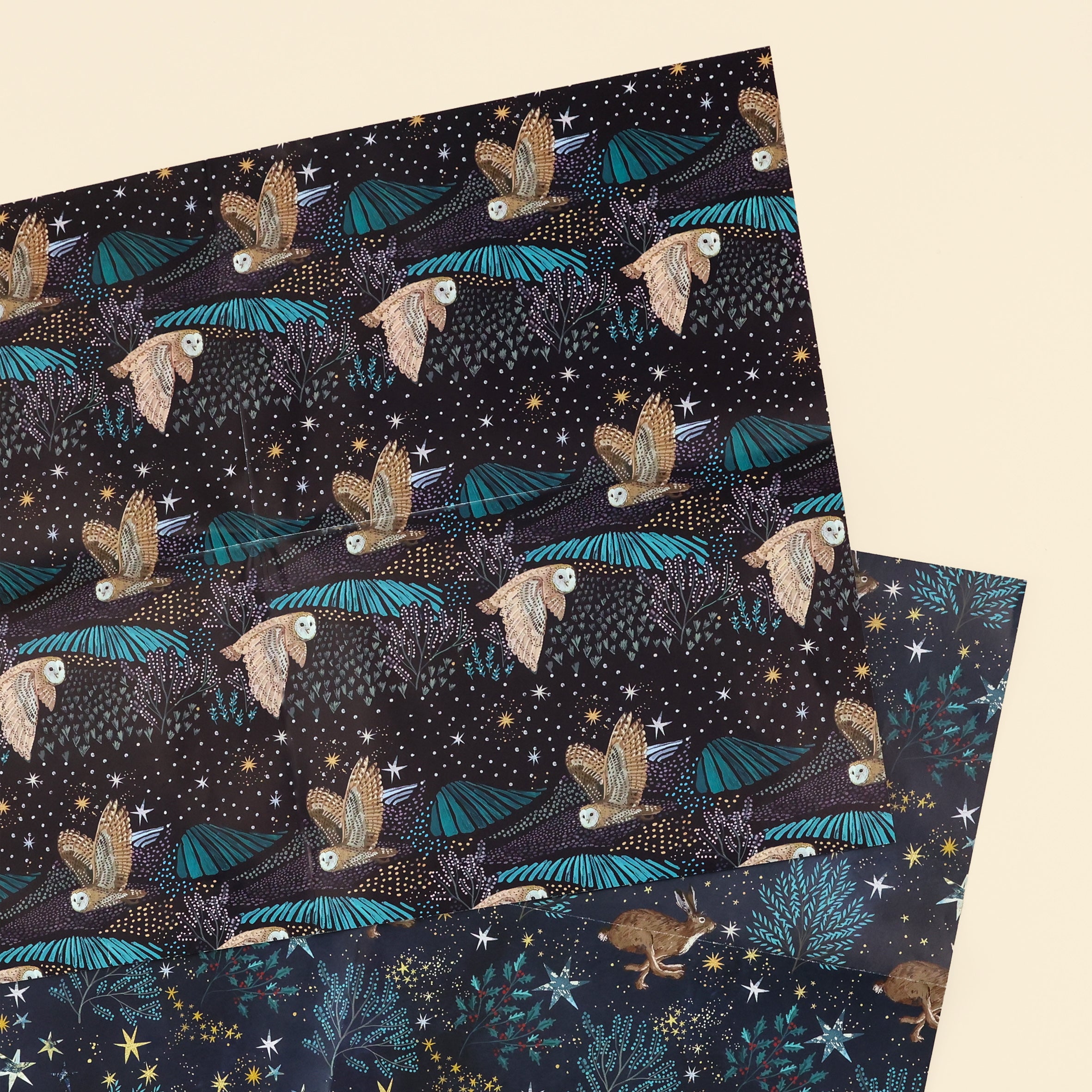 Close up of both designs of wrapping paper; one with starlit fields on black paper with a barn owl flying in a star lit night and the other; hare running amongst trees with stars on navy background. 