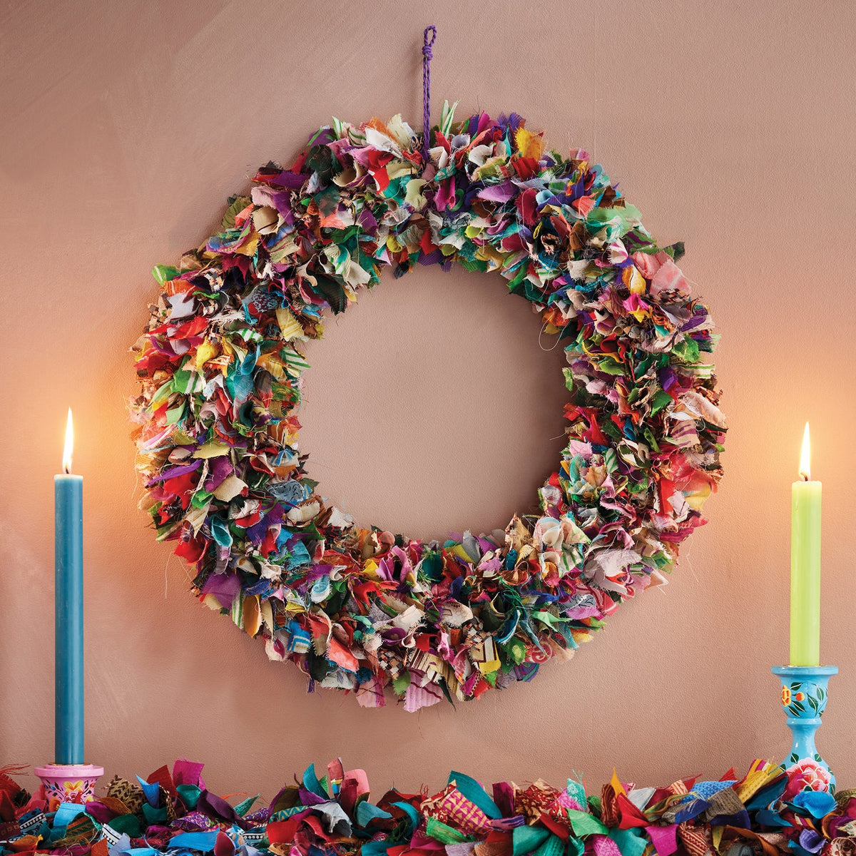 Large recycled sari wreath in multicolour hanging from a wall besides to lit dinner candles and matching recycled sari garland in multicolour