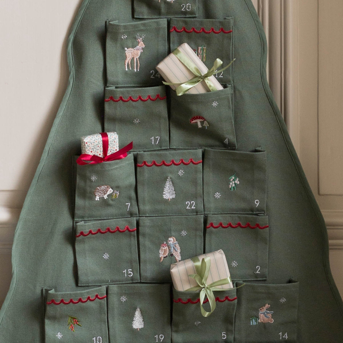 Close up of advent calendar, showing embroidered numbers, reindeer, hedgehog, owls, rabbit, trees and snow flakes, hanging on wall with presents in pockets 