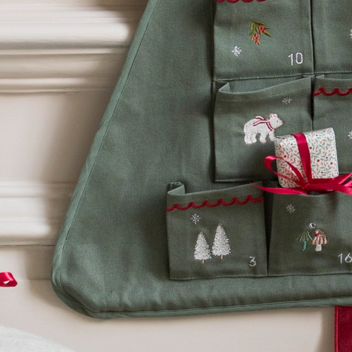 Close up of bottom left corner of Advent showing the wooden bar holding the shape of the tree, with embroidered numbers, polar bear, trees and mushrooms with ed ribbon trim on pockets