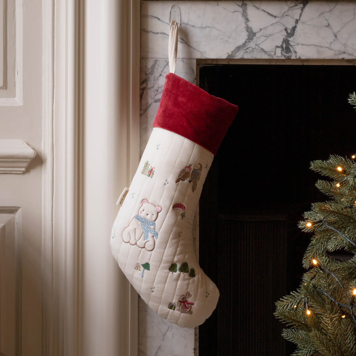 Polar Bear embroidered cotton Christmas stocking with red velvet trim, hanging in fireplace next to lit Christmas tree