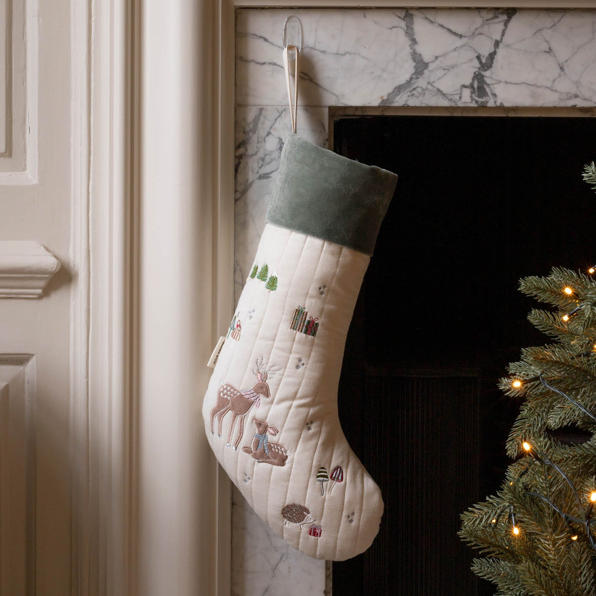 Reindeer embroidered cotton Christmas stocking with green velvet trim, hanging from fireplace