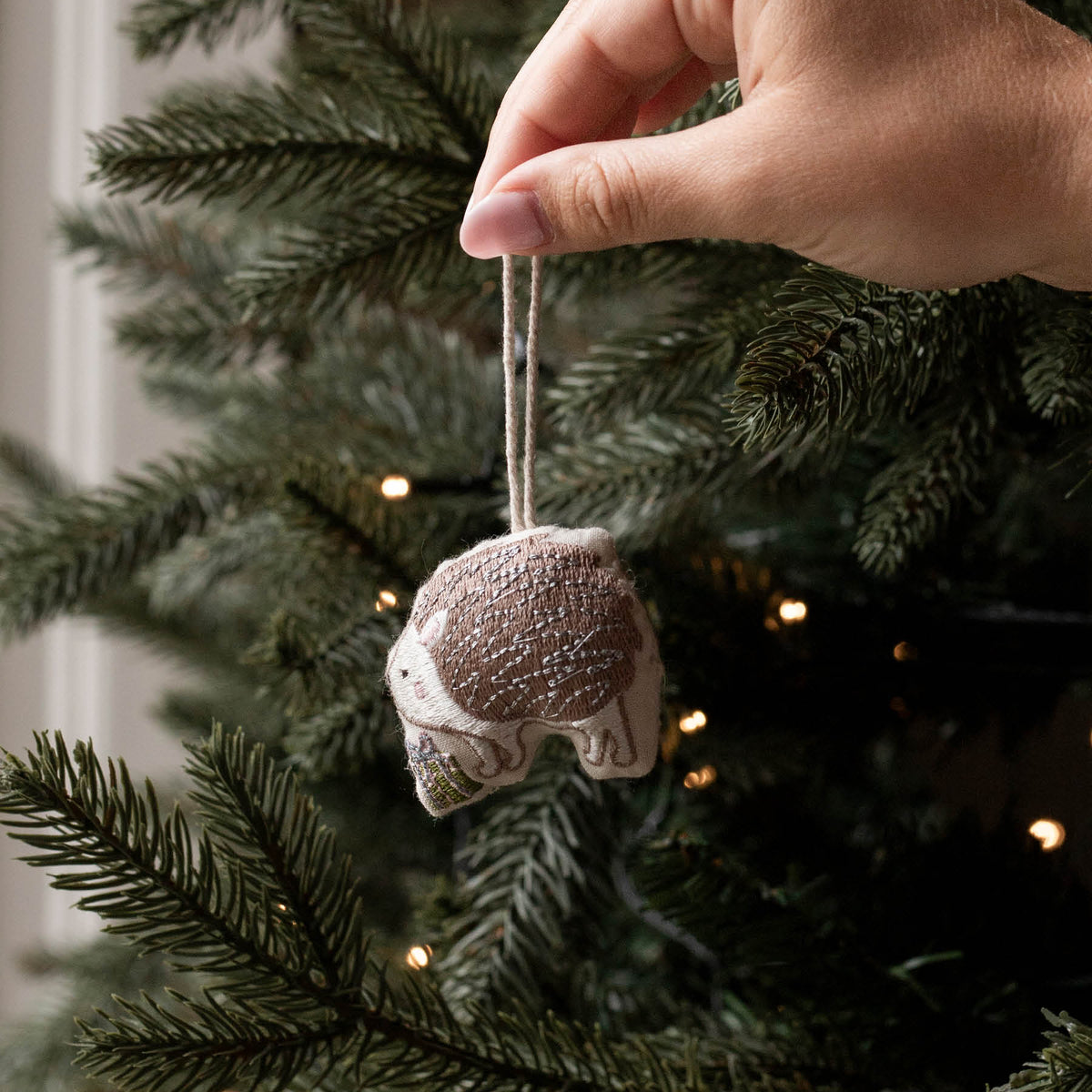 Cotton embroidered hedgehog Christmas decoration being held in front of a lit Christmas tree